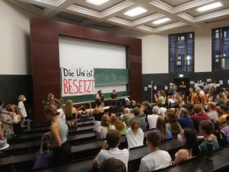 Studierene sitzem im Hörsaal. An der Wand hängt ein Banner "Die Uni ist besetzt"