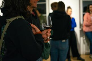 Ein Foto von einer Afterparty oder einem geselligen Empfang. Im Fokus der Nahaufnahme steht eine Person im schwarzen Kapuzenpullover, die ein Weinglas mit Rotwein in der Hand hält. Im leicht unscharfen Hintergrund unterhalten sich weitere Gäste in kleinen Gruppen in einem hell erleuchteten Innenraum.