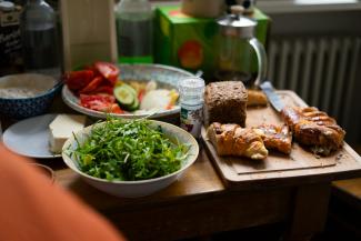 Ein Foto von einem reich gedeckten Frühstücksbuffet beim Foodsharing. Auf einem Holztisch stehen eine große Schüssel mit frischem Rucola-Salat, ein Teller mit geschnittenen Tomaten und Gurken sowie ein Holzbrett mit verschiedenen Backwaren wie Croissants, Brot und Plundergebäck. Im leicht unscharfen Hintergrund sind Gewürze, Aufstriche, Getränkeflaschen und eine Kaffeepresse angeordnet.